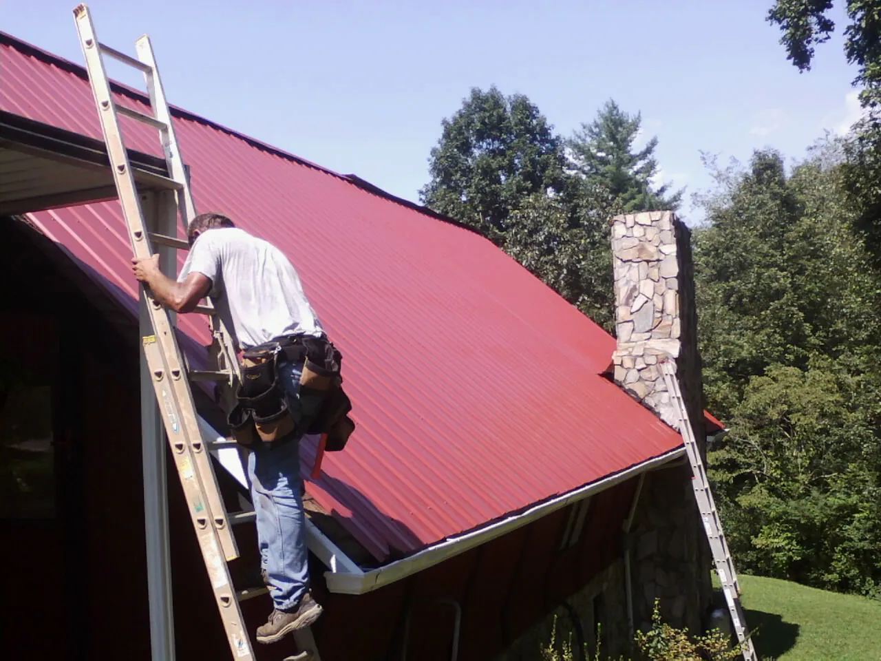 Roofer on a ladder beside a red metal roof and stone chimney — Soffit & Fascia Repair by Roofing Brothers in Harpster